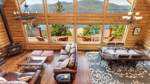 A cozy log cabin interior with a fireplace, a sofa, a dining table, and large windows offering a view of the mountains and lake. - Ski, Swim, Relax at Whitefish Lake | Stunning Views of Whitefish Lake (Whitefish)