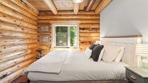 A log cabin bedroom with a large bed, a window, and a wooden ceiling. - Ski, Swim, Relax at Whitefish Lake | Stunning Views of Whitefish Lake (Whitefish)