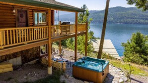 A wooden cabin with a balcony overlooking a lake, a hot tub, and a stone patio area. - Ski, Swim, Relax at Whitefish Lake | Stunning Views of Whitefish Lake (Whitefish)