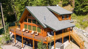 A log cabin with a deck, large windows, and a stone foundation. - Ski, Swim, Relax at Whitefish Lake | Stunning Views of Whitefish Lake (Whitefish)