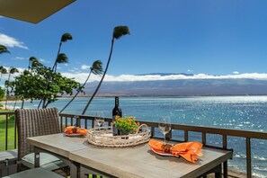 Une table à manger avec une bouteille de vin, des verres et un panier de fleurs, donnant sur un paysage marin magnifique, avec des palmiers en vue.