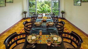 Une longue table à manger, dressée avec des plats traditionnels indiens, est entourée de chaises en bois aux motifs complexes, dans une pièce dotée de grandes fenêtres offrant une vue sur une végétation luxuriante.