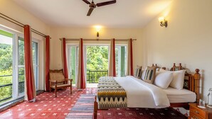 Une chambre à coucher comprenant un lit, une chaise, une table de chevet, une commode, un ventilateur de plafond et une vue sur la verdure par les fenêtres.