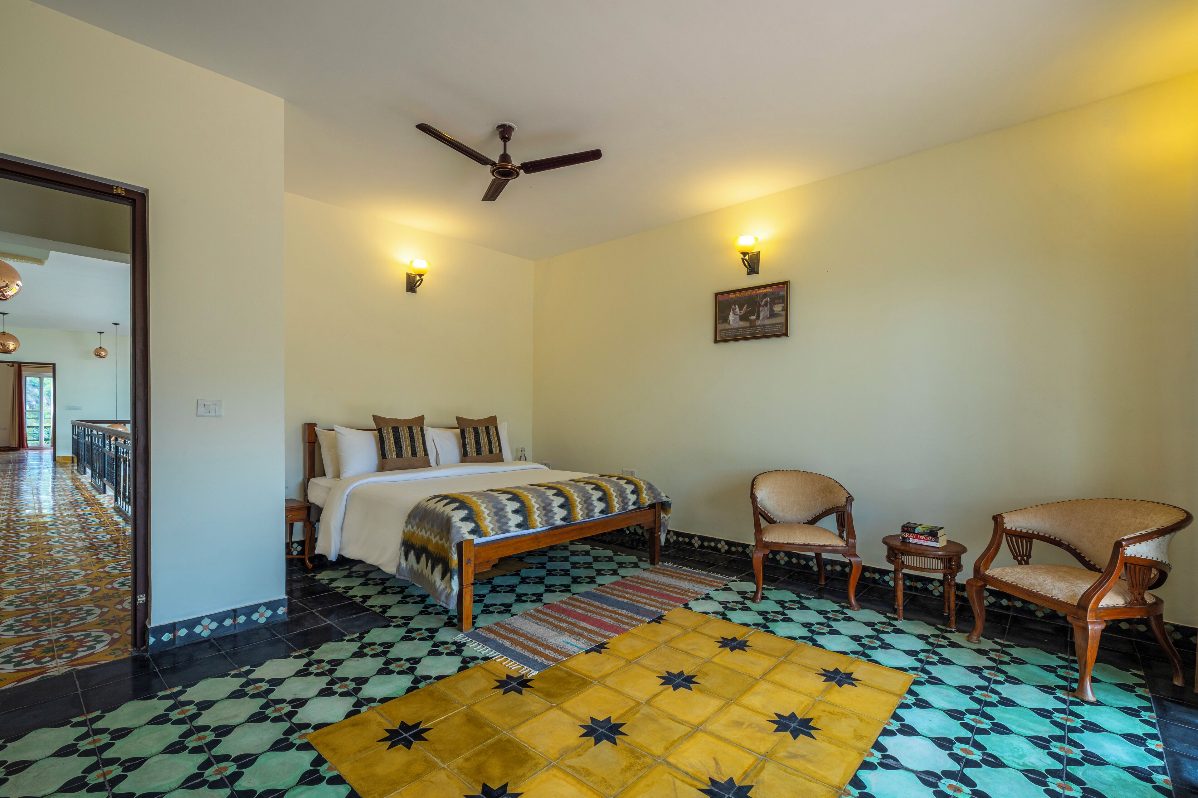 A bedroom with a bed, two chairs, a small table, a ceiling fan, and patterned floor tiles.