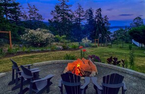 A fire pit surrounded by Adirondack chairs in a landscaped garden with a view of trees and a body of water. - Water View Modern Farmhouse in West Sound (Eastsound)