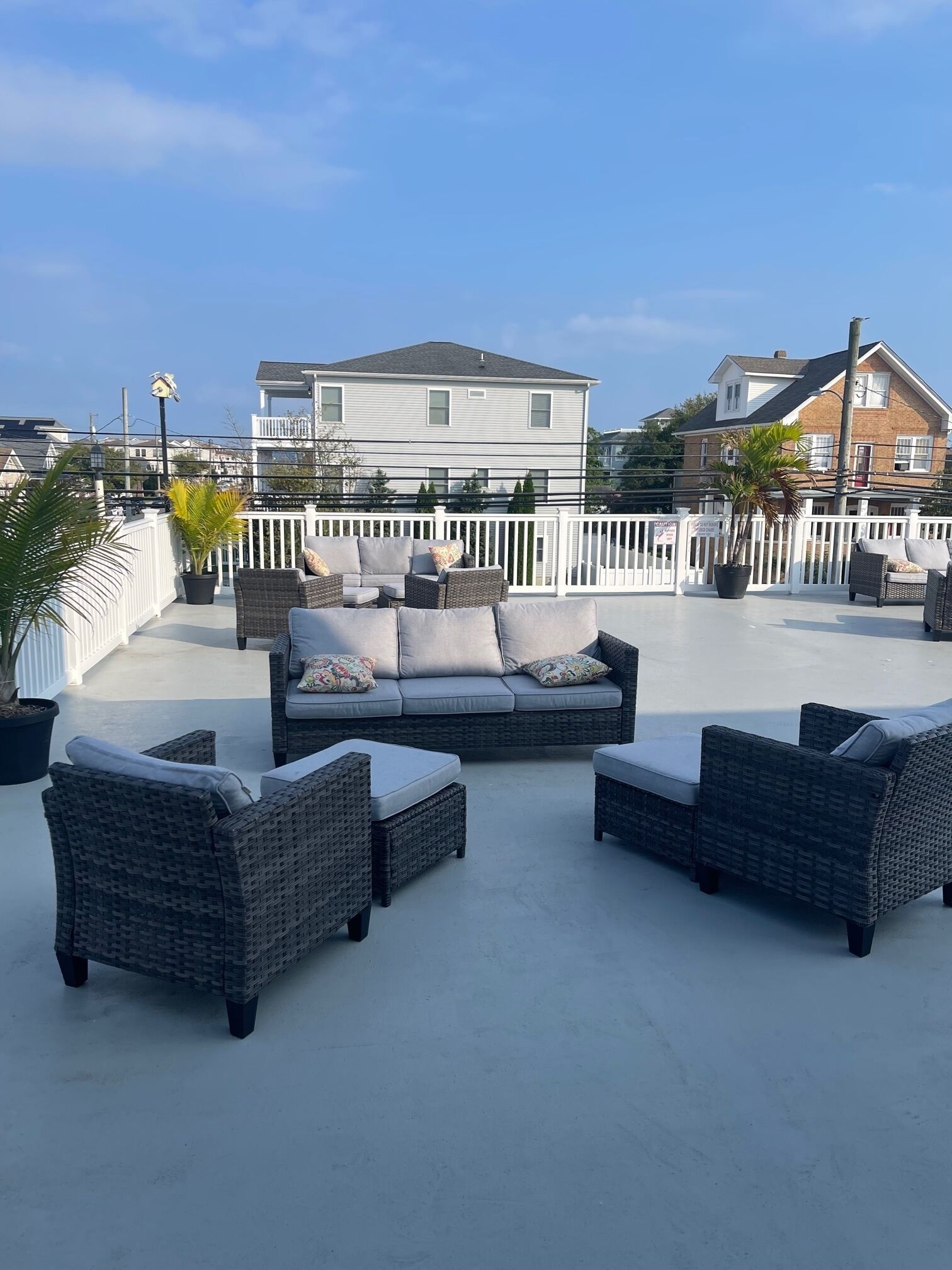 A rooftop patio with wicker furniture, a white fence, and houses in the background.