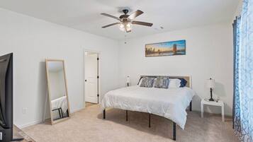 A bedroom with a bed, a ceiling fan, a painting, a mirror, a television, and a remote control.