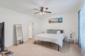 A bedroom with a bed, a ceiling fan, a painting, a mirror, a television, and a remote control.