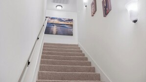 A staircase with carpeted steps leading to a landing with a painting and wall-mounted lights. - Peaceful Bayou Escape Close to LSU Campus - CL5626 (Baton Rouge)