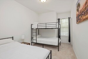 Una habitación con una cama litera, una cama individual, una mesita de noche con lámpara y un ventanal con cortinas.