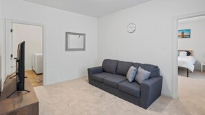 A living room with a grey sofa, a flat-screen TV, a clock, and a framed picture on the wall. - Peaceful Bayou Escape Close to LSU Campus - CL5626 (Baton Rouge)