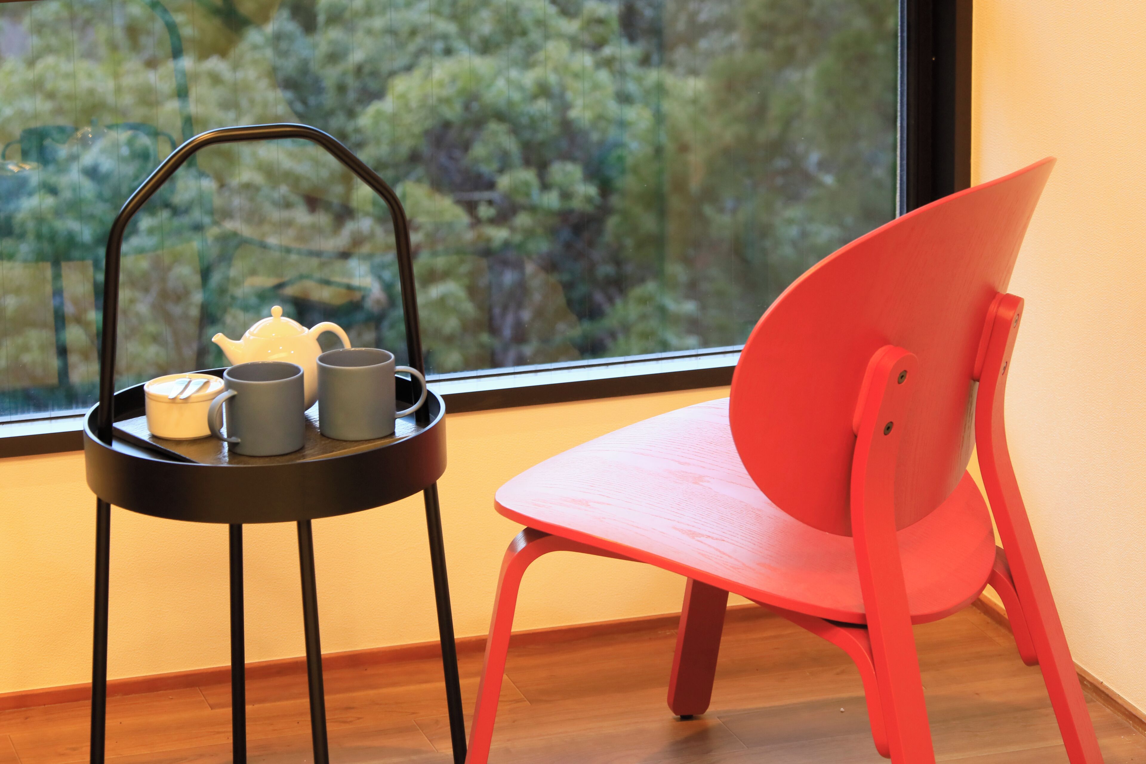 A room with a window, a red chair, and a black table with a tray holding a teapot, cups, and a bowl.