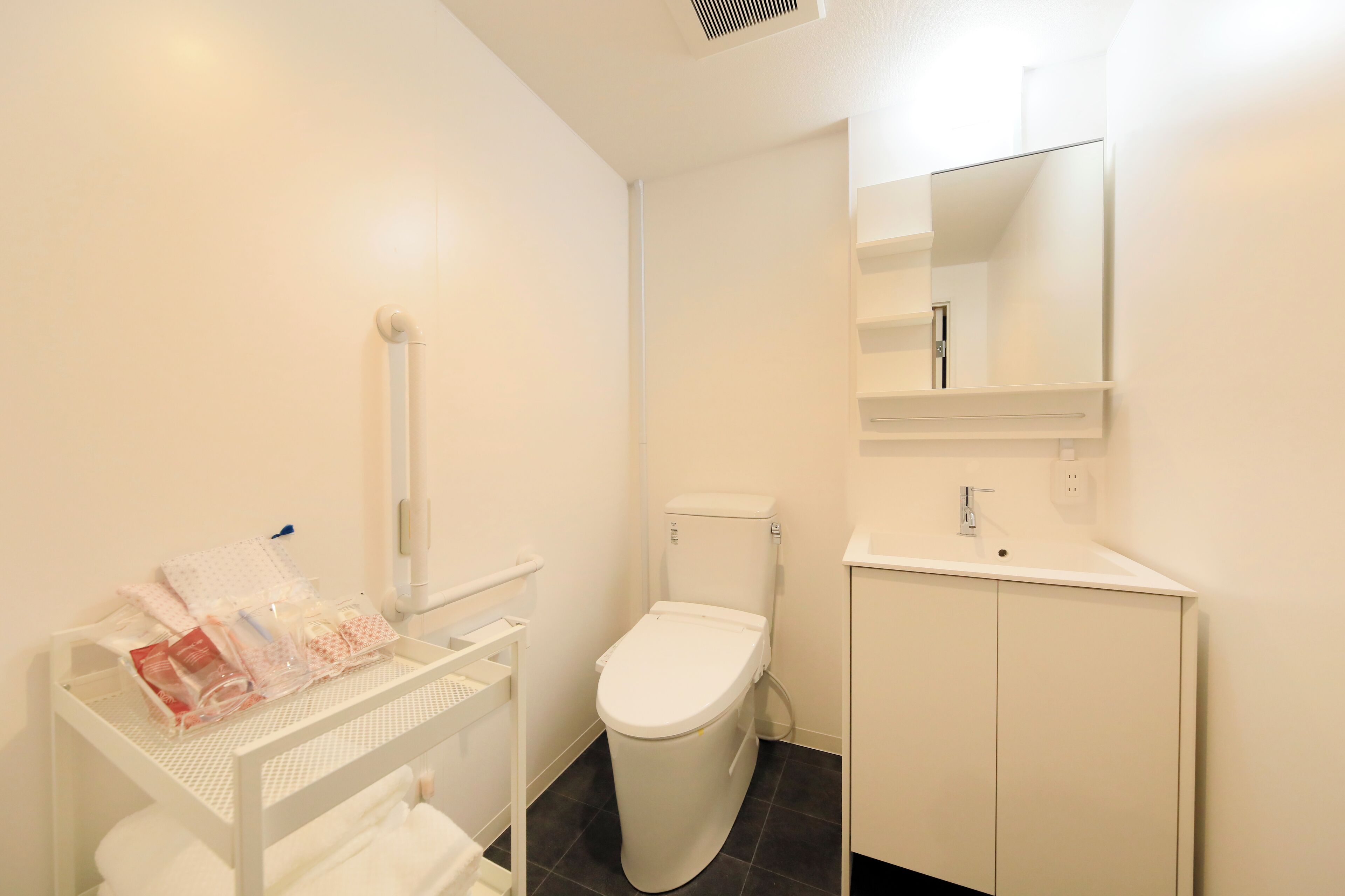 Un baño moderno con inodoro, lavamanos y espejo. Hay un estante blanco con artículos de aseo y un armario blanco.