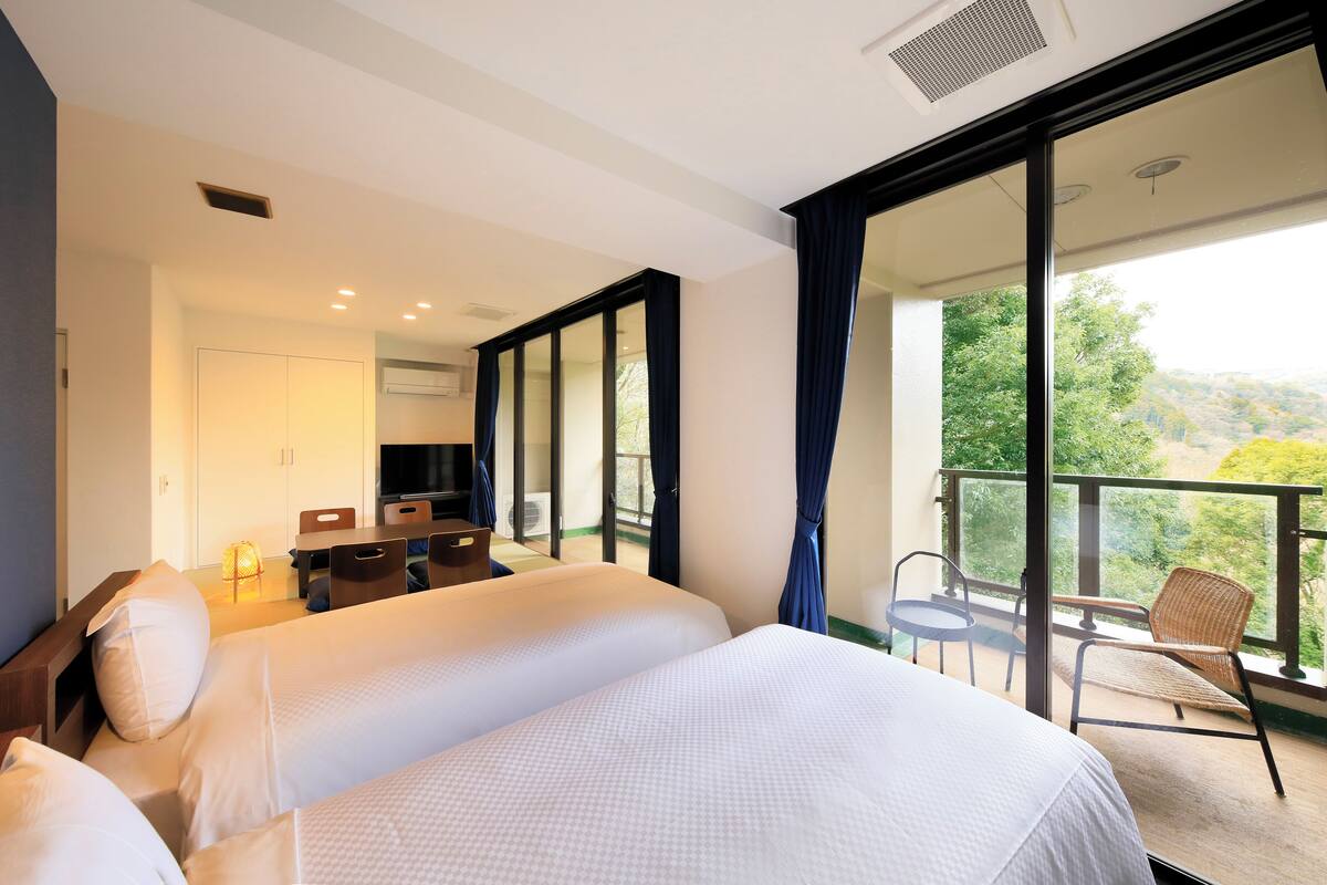 A hotel room with two beds, a desk with a chair, a balcony with a chair, and a view of greenery.