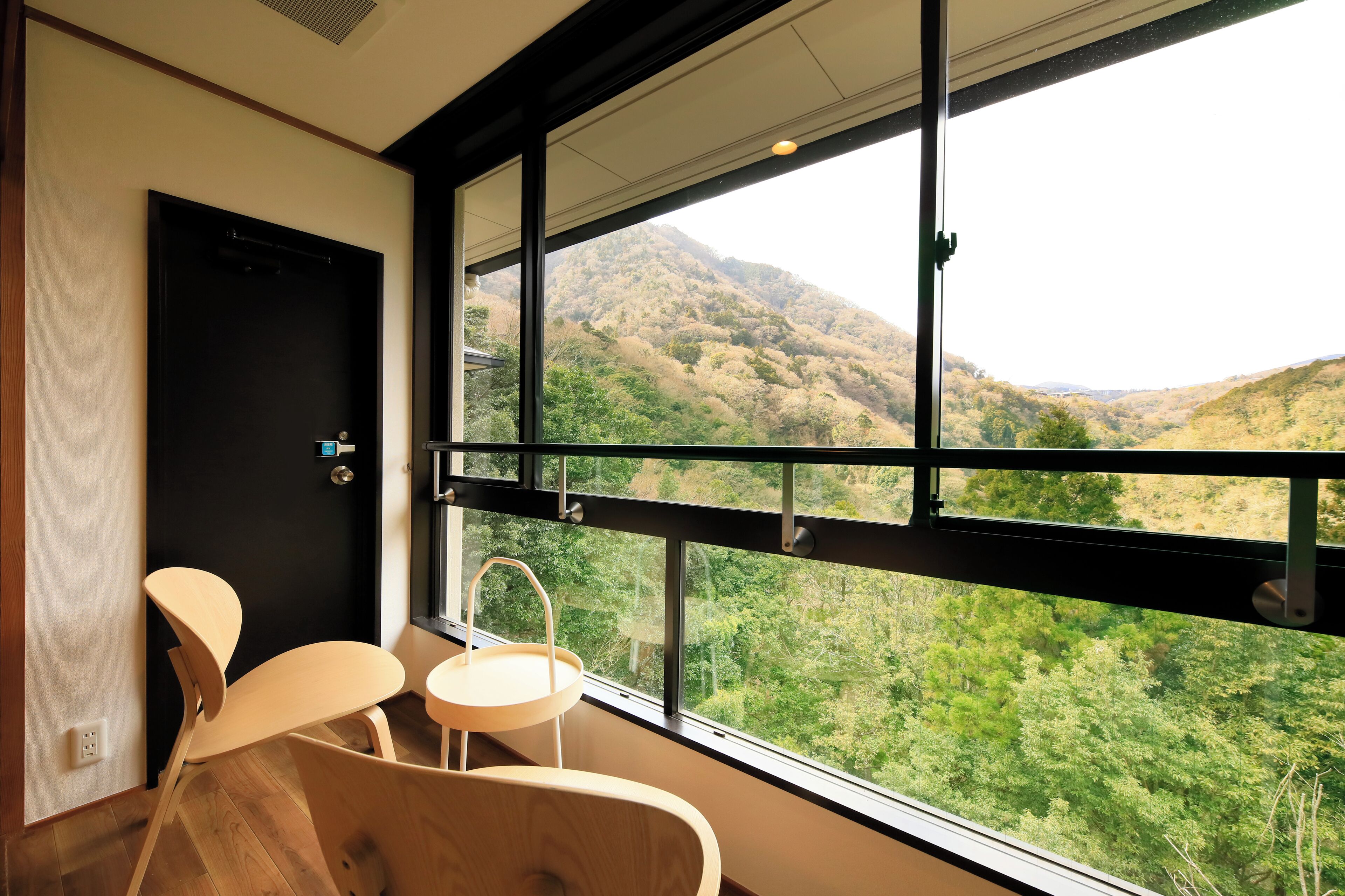 A room with a large window offering a view of a mountainous landscape. There is a door and a small table with a chair.