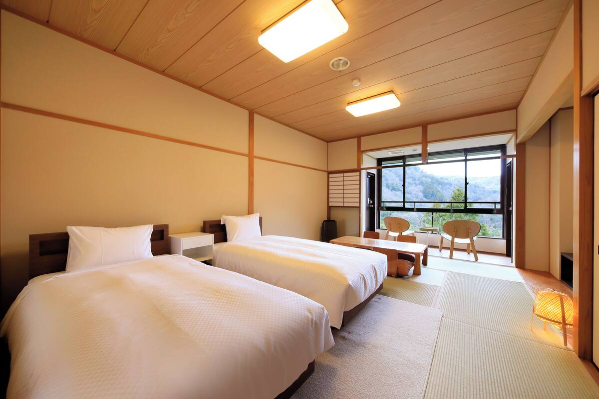 A hotel room with two beds, a small table, chairs, a sofa, and a large window offering a view of the outdoors.