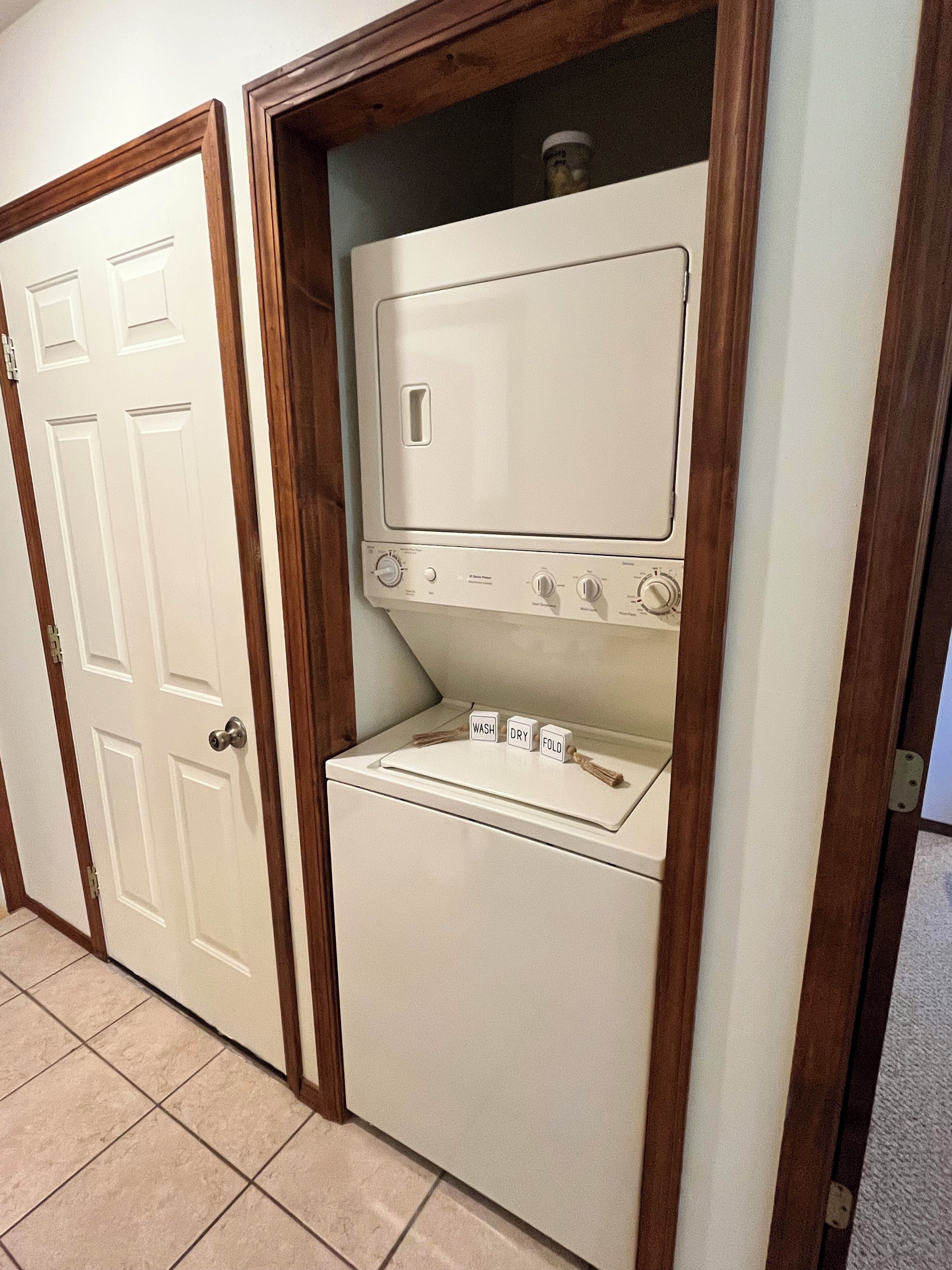 A compact, wall-mounted washer and dryer unit with a wooden frame, a control panel with dials, and a small shelf with a jar.
