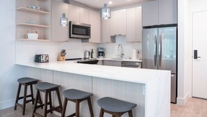 A modern kitchen with a white countertop, stainless steel appliances, wooden stools, and pendant lighting.