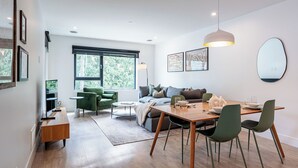 A modern living room with a wooden dining table, green chairs, a grey sofa, and a large window.