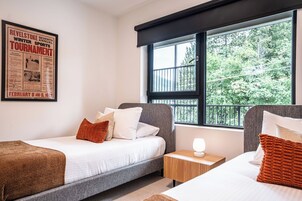 Habitación de hotel con dos camas individuales, una mesita de noche con lámpara, un cartel en la pared y una ventana con vistas a árboles.