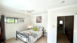 A bedroom with a bed, bedside tables, a ceiling fan, an air conditioning unit, a window with curtains, and a door leading to another room.