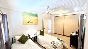 A hotel room with two beds, a ceiling fan, a painting, a wardrobe, and a desk with a lamp.