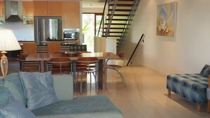 A modern kitchen with stainless steel appliances, a dining area with a table and chairs, a staircase, and a living room with a sofa and a patterned rug. - BHA 1 Beachfront Haven fullhouse (Mission Beach)