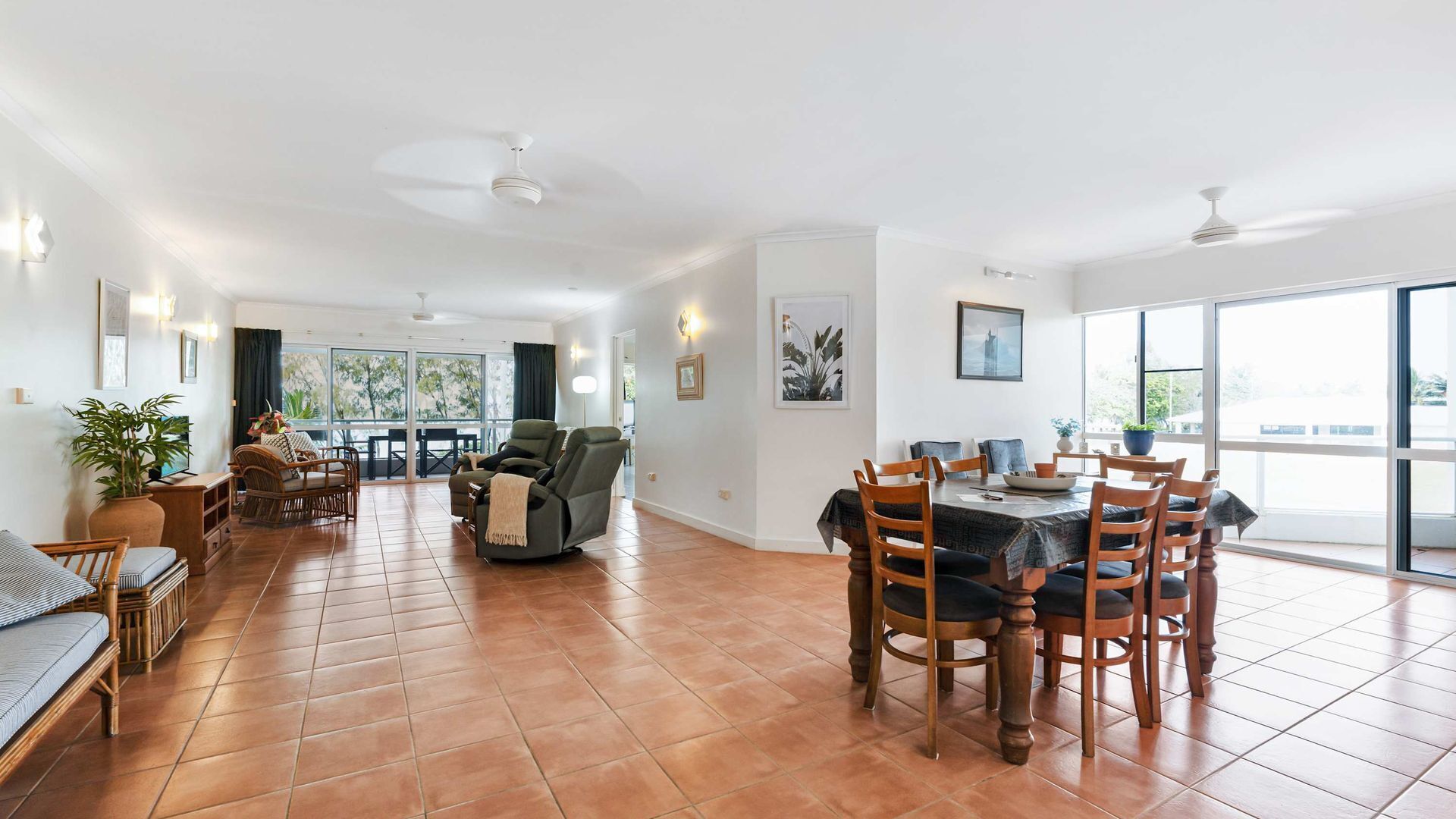 A spacious living area with a dining table, chairs, and a reclining chair. There are large windows providing natural light and a view of the outdoors.