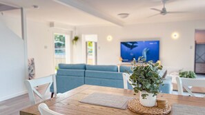 A modern living room with a blue sofa, a dining table with a placemat, a potted plant, and a television displaying an image of a whale. - Bella Luna Beach Front Full house (South Mission Beach)
