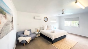 Un dormitorio con cama, mesita de noche, lámpara, espejo en la pared, ventilador de techo y ventana.