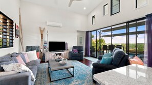 A modern living room with a flat-screen TV, a sofa, a coffee table, and a dining area with a view of the outdoors.