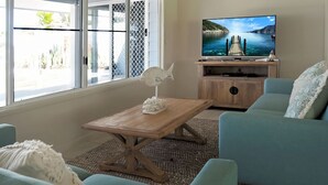 A living room with a wooden coffee table, a TV on a wooden stand, a light blue sofa, and a view of the sea through a window. - Solace Dream Beachfront Escape (South Mission Beach)