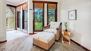 Una habitación con un sofá y reposapiés de mimbre, una mesita de madera con una planta y vistas amplias a través de grandes ventanales.