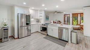 A modern kitchen with white cabinetry, stainless steel appliances, and a dining area with wicker furniture. - South Kauai Tranquil Retreat w/ AC in Poipu (Koloa)
