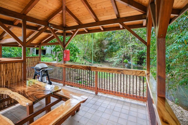 Un espacio al aire libre techado con bancos de madera y una parrilla, rodeado por una valla de bambú y vistas a un paisaje natural.