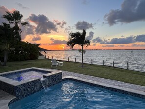 Ein Swimmingpool mit Blick auf den Sonnenuntergang über dem Wasser, umgeben von Gras und Palmen.