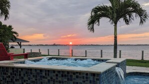 A hot tub with a view of the sunset over a body of water, surrounded by a stone patio and palm trees.