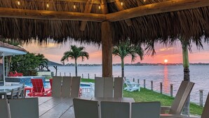 A covered outdoor seating area with chairs and tables, overlooking a body of water at sunset.