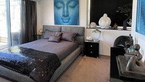 Une chambre moderne avec un grand lit, une tête de lit, une table de chevet, une commode, une étagère murale agrémentée d’objets décoratifs et une grande fenêtre avec des rideaux.