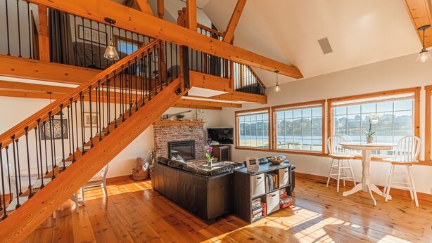 Un vaste espace de vie agrémenté d’un escalier en bois, d’une cheminée, d’une grande fenêtre offrant une vue dégagée et d’un coin repas avec des chaises blanches.