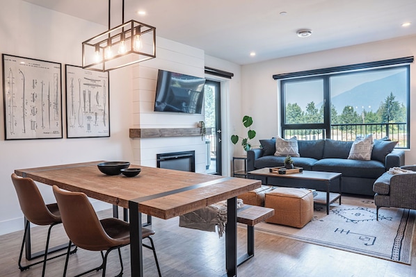 Una sala de estar moderna con una mesa de comedor de madera, asientos cómodos, un televisor grande y vistas al exterior.