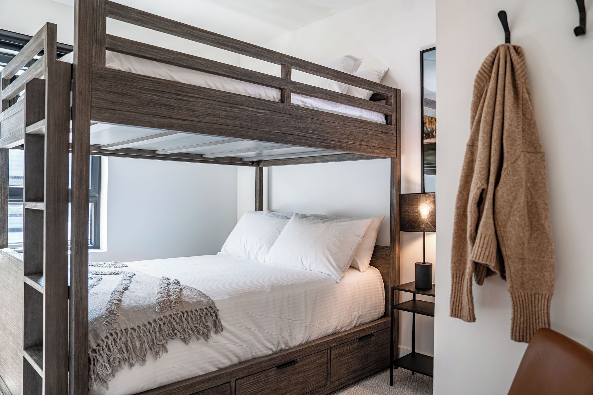 A bunk bed with a wooden frame, a bedside table with a lamp, and a coat hanging on the wall.