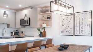 A modern kitchen with white cabinets, a microwave, a coffee maker, and a dining area with a wooden table and chairs. - Glades and Green by Revelstoke Vacations (Revelstoke)