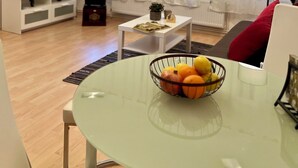Un soggiorno moderno con un tavolo in vetro, una ciotola di frutta, un divano bianco, una televisione e un'ampia finestra con tende leggere.