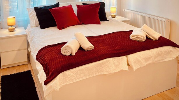 Un letto rifatto con cuscini rossi e neri, una testiera bianca e una coperta rossa. Si vede un comodino bianco con una lampada, un tappeto nero e un quadro incorniciato appeso al muro.