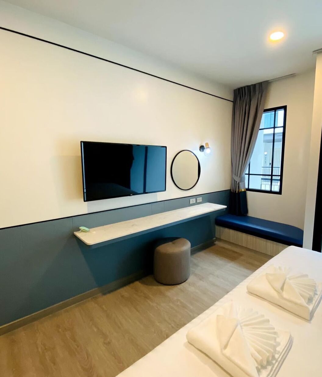 A modern hotel room with a flat-screen TV, a round mirror, a bedside table, a bench, and neatly folded towels.