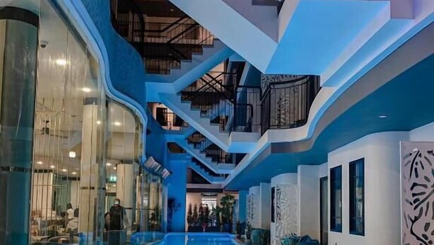 A modern indoor swimming pool with a glass wall on one side, a staircase with a railing on the other, and a ceiling with a geometric design. - Poolrada Boutique Hotel (Thep Krasattri)