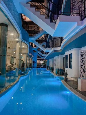A modern indoor swimming pool with a glass wall on one side, a staircase with a railing on the other, and a ceiling with a geometric design. - Poolrada Boutique Hotel (Thep Krasattri)