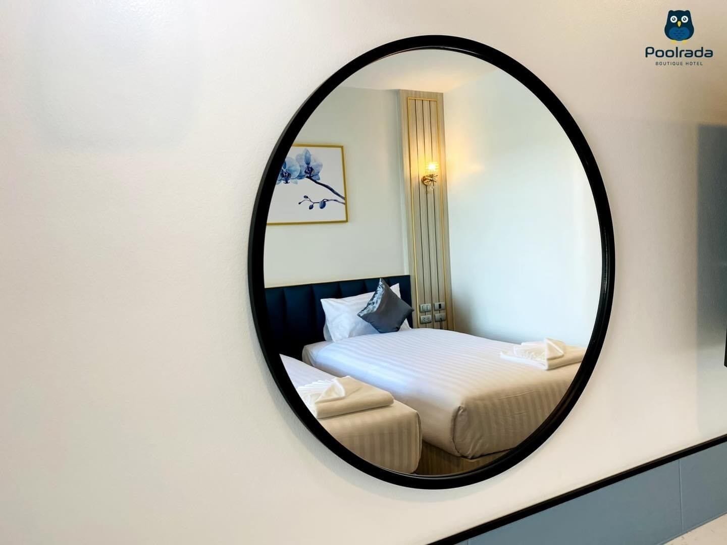 Una habitación de hotel con un espejo redondo, una cama con ropa blanca, un cabecero, una mesita de noche, una lámpara de pared y un cuadro enmarcado.