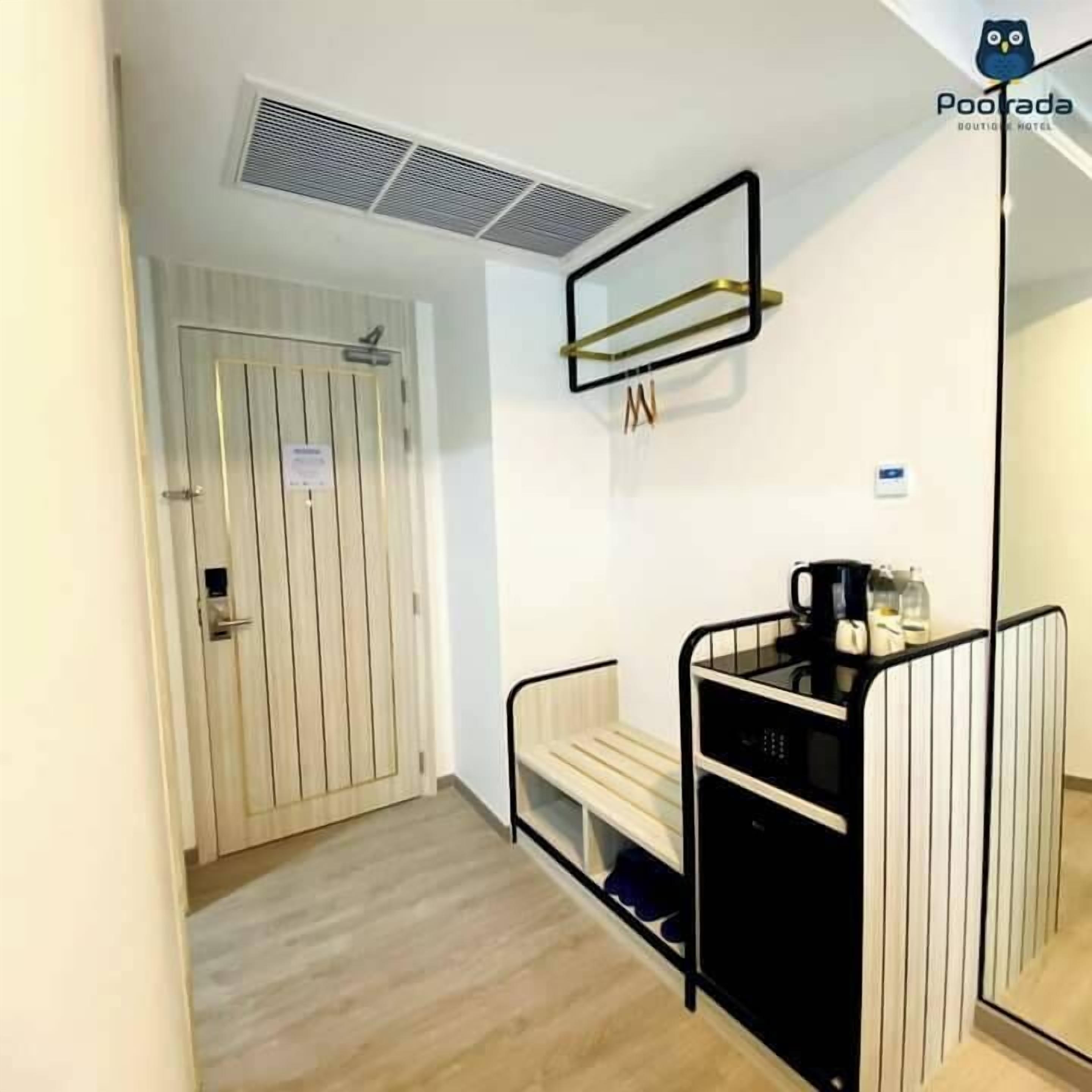 Habitación de hotel con puerta de madera, un pequeño banco, estructura de cama de metal negro, mesita de noche con hervidor y espejo en la pared.